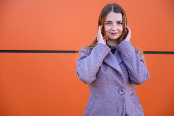 Photo of a cute smiling posing blonde Caucasian girl with long beautiful hair outdoors in a pink coat on an orange background in the afternoon on a sunny day. - Foto, immagini