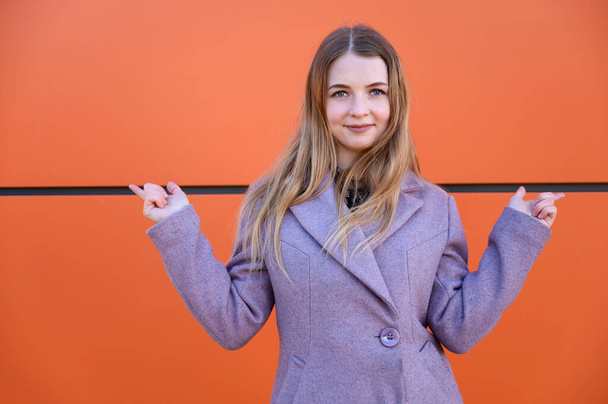 Photo of a cute smiling posing blonde Caucasian girl with long beautiful hair outdoors in a pink coat on an orange background in the afternoon on a sunny day. Model points fingers to the sides. - Foto, immagini