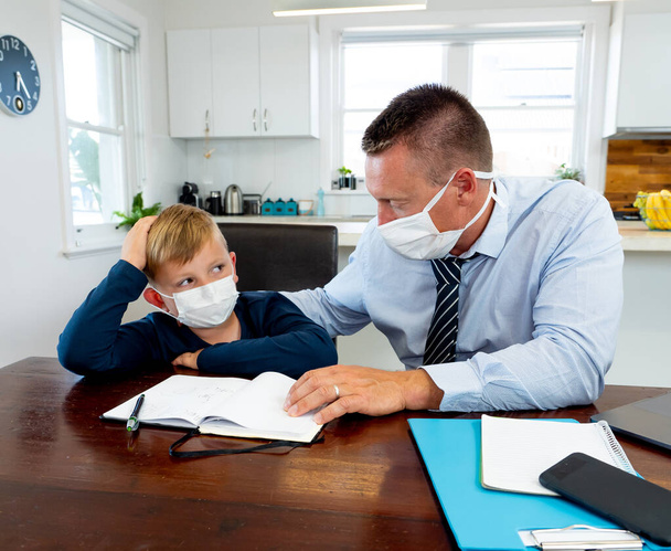 Coronavirus Outbreak school shutdowns. Stressed parent coping with remote work and homeschooling worried about COVID-19 pandemic. Father and son with mask in quarantine working and learning from home. - Photo, Image
