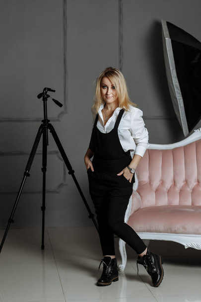 Beautiful girl in a white shirt and black overalls on a pink sofa in a photostudio on fashion photography - Fotó, kép