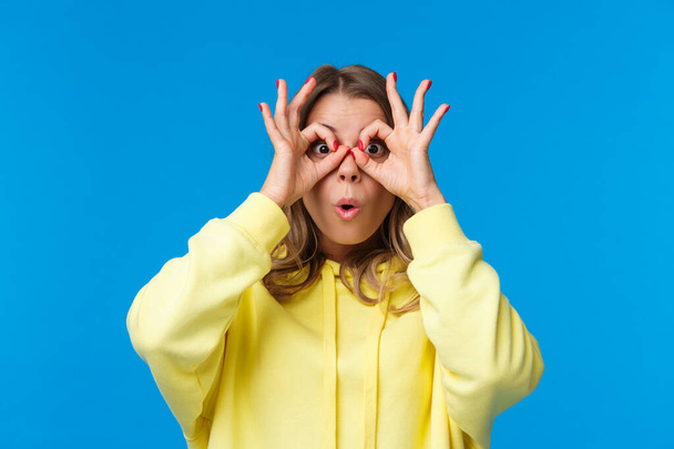 Impressed and intrigued, curious young caucasian girl in yellow hoodie looking at something awesome, found great product special discount, make glasses with fingers on eyes, blue background - Фото, изображение
