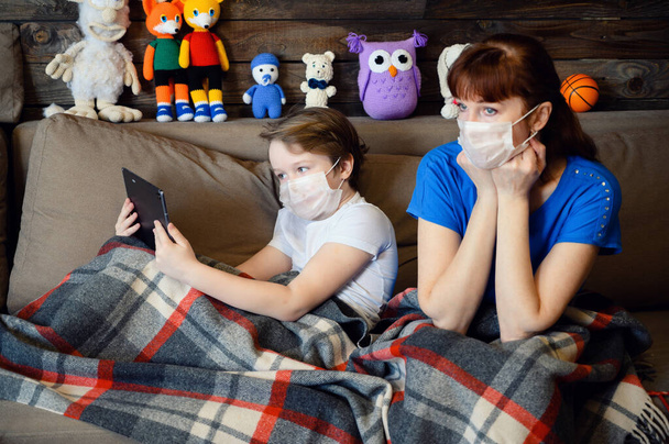 Photo in the home interior of a 9 years old boy with mom lies on the sofa in a medical mask with a phone tablet in hands in quarantine - 写真・画像