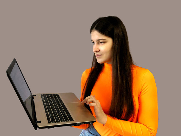 Young beautiful girl with dark hair in a bright orange sweater on a gray background. With a laptop in hand - Photo, Image