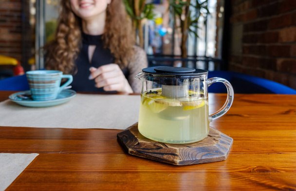 A kettle of tea made of herbs is on the table and a smiling girl sits in the background with a cup. - Fotografie, Obrázek