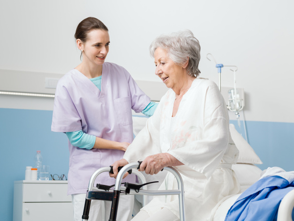 Enfermeira profissional sorridente assistindo um paciente sênior no hospital, a mulher está se apoiando em um andador
 - Foto, Imagem