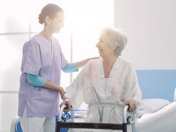 Smiling professional nurse assisting a senior patient at the hospital, the woman is leaning on a walker - Фото, зображення