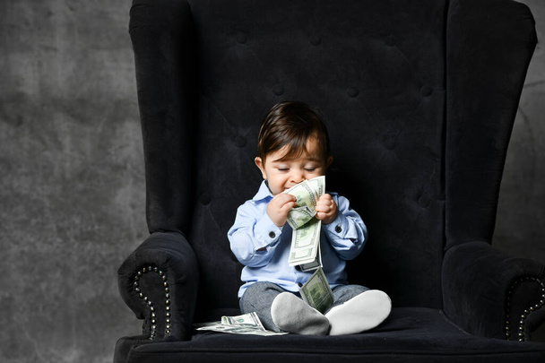 Baby boy in blue shirt, gray pants, white booties. He holding some cash, sitting in black armchair on gray background. Close up - Foto, Bild