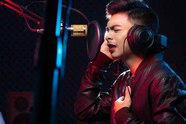 Ázsiai tinédzser férfi fekete haj fülbevaló fejhallgató énekelni egy dalt hangosan teljesítmény hangot lógó mikrofon kondenzátor, hangszórók, zeneszerző jegyzetfüzet és monitor. Studio Sound Proof elnyelő faliszoba - Fotó, kép