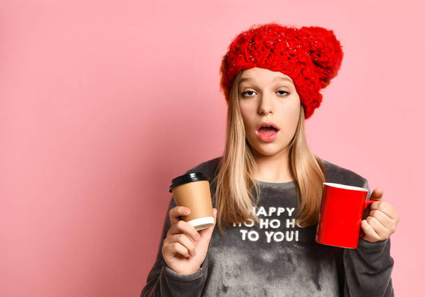 Girl in gray reindeer pajamas, red hat. She holding ceramic and paper cups, looking sleepy, posing on pink background. Close up - Fotografie, Obrázek