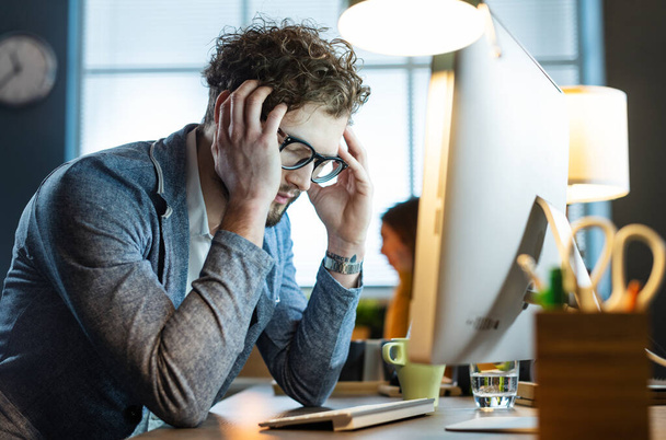 Stresszes irodai dolgozó ül az asztalnál, és dolgozik a számítógéppel, fáradt és fáj a feje - Fotó, kép