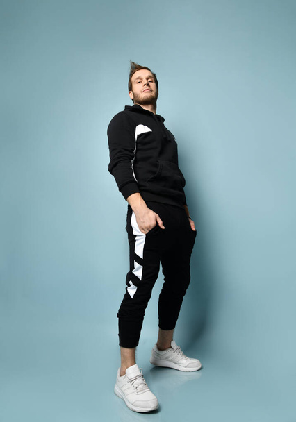 Hipster male in black tracksuit, bracelet and sneakers. He is rolling up his sleeve, posing against blue background. Full length - Fotó, kép