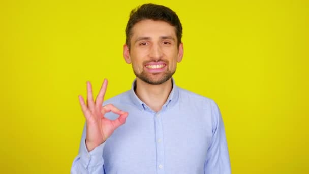 Młody mężczyzna w jasnoniebieskiej koszuli uśmiecha się i pokazuje OK - Materiał filmowy, wideo
