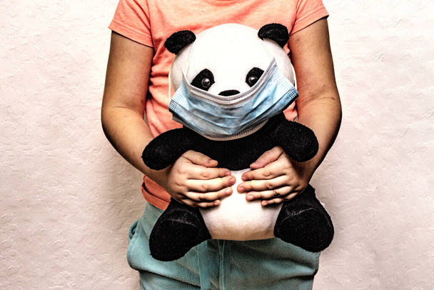 Panda de juguete suave en una máscara médica desechable en las manos de los niños. Concepto epidémico de Coronavirus. Higiene personal para niños. Fondo de masilla blanca
 - Foto, imagen