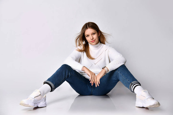 Teenage female in jeans, sweater, socks and sneakers. Smiling, sitting on floor, legs wide apart, isolated on white. Close up - Φωτογραφία, εικόνα