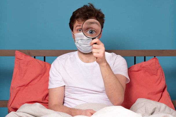 Jovem engraçado em máscara médica olhando através de lupa sentado na cama durante a quarentena, isolado em branco
 - Foto, Imagem