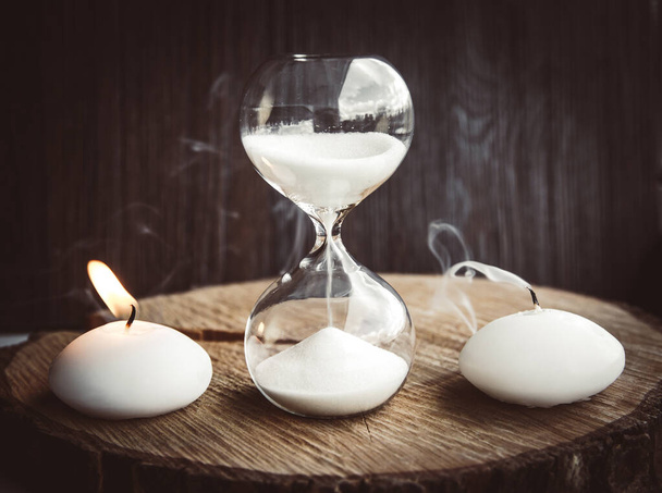 Time running out, life and death concept. One candle burning with flame as living other is blown away as dead and hourglass measures lifetime.  - Photo, Image