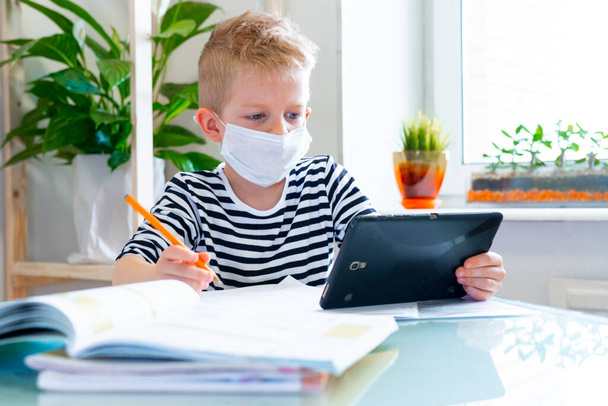 Distance learning online education. Sickness schoolboy in medical mask studying at home with digital tablet in hand and doing school homework. Training books and notebooks on table. - Valokuva, kuva