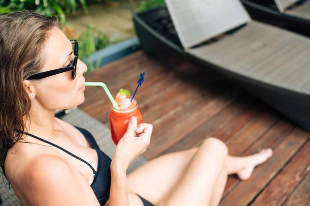 Siyah bikinili güzel bir kadın havuz kenarında güneşlenirken kokteyl içiyor. - Fotoğraf, Görsel