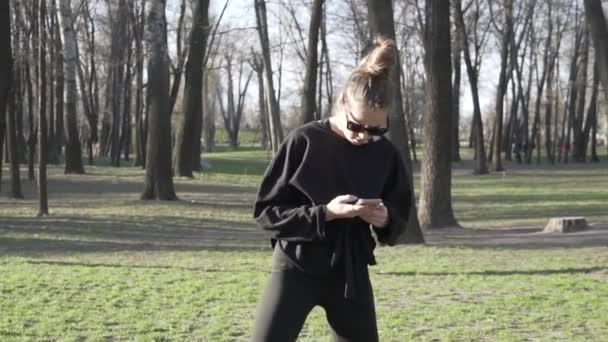 Junge Frauen trainieren vor dem Fitnesstraining im Park. Gesunde junge Frau wärmt sich draußen auf, abgelenkt durch die Korrespondenz in sozialen Netzwerken mit dem Smartphone. Frau macht Dehnübungen - Filmmaterial, Video