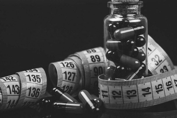 Крупним планом скляну пляшку з дієтичними таблетками та вимірювальною стрічкою ізольовано на чорному тлі. Чорно-біле фото
. - Фото, зображення