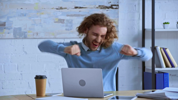 χαρούμενος σγουρός άντρας χορεύει κοντά στο λάπτοπ στο γραφείο  - Πλάνα, βίντεο