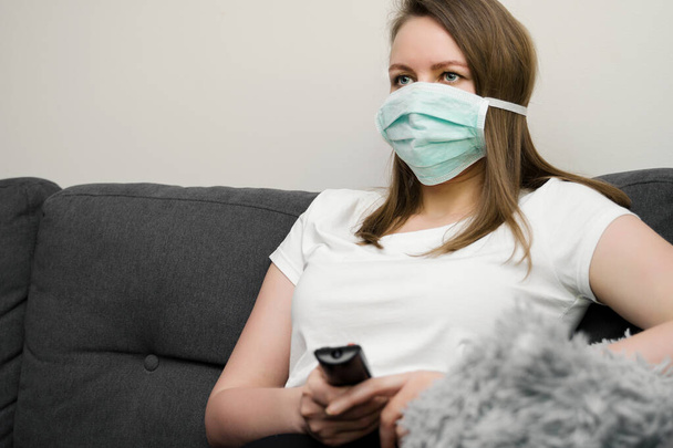 Woman in medical mask watching TV. Quarantine at home. - Photo, image