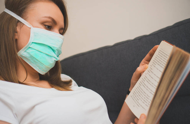 Woman in medical mask reading book. Isolation at home. - Foto, afbeelding