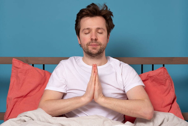 Entspannter unbeschwerter Mann meditiert zu Hause auf Quarantäne, am Bett sitzend, versucht zu entspannen und gerät nicht in Panik. Menschen, Ruhe, Entspannung, Meditation, Lifestylekonzept - Foto, Bild