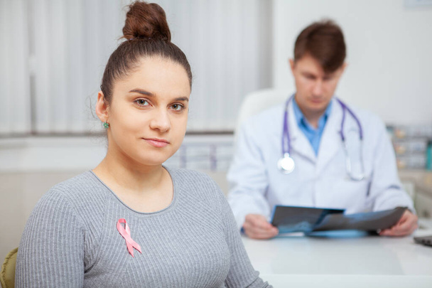 Viehättävä yong nainen rintasyöpä vaaleanpunainen nauha rinnassa katsellen kameraan luottavaisesti, lääkäri tutkii röntgenkuvat taustalla
 - Valokuva, kuva