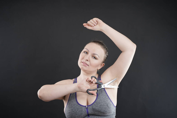 plus size woman shows hairy armpit. the girl holds a pair of scissors in her hand and experiences negative surprised emotions, hating excessive body hair . Studio black monochrome background - Photo, image