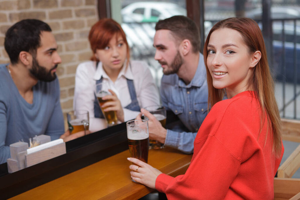 Attraente giovane donna sorridente alla fotocamera mentre beve birra al pub con i suoi amici. Festa, concetto di celebrazione
 - Foto, immagini