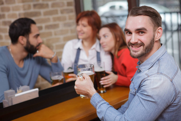 Όμορφος γενειοφόρος άντρας χαμογελάει στην κάμερα, πίνοντας μπύρα με τους φίλους του στην παμπ. - Φωτογραφία, εικόνα