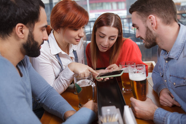 Group of friends using smart phone while drinking beer together. Young beautiful woman showing something online to her friends, using her phone - Photo, Image