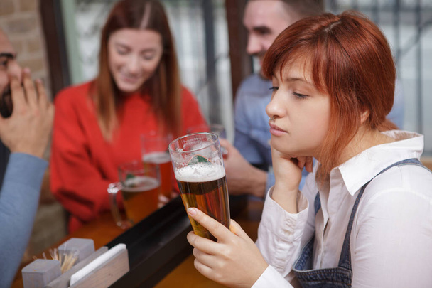 Nuori nainen näyttää järkyttyneeltä, juo olutta, kun hänen naisystävänsä flirttailee lääkärin kanssa taustalla. Mustasukkaisuus, yksinäisyys käsite
 - Valokuva, kuva