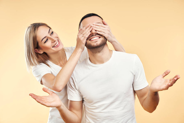 Guess who? Surprise for him. Beautiful young woman covering eyes of her boyfriend isolated on yellow background - Foto, Imagen