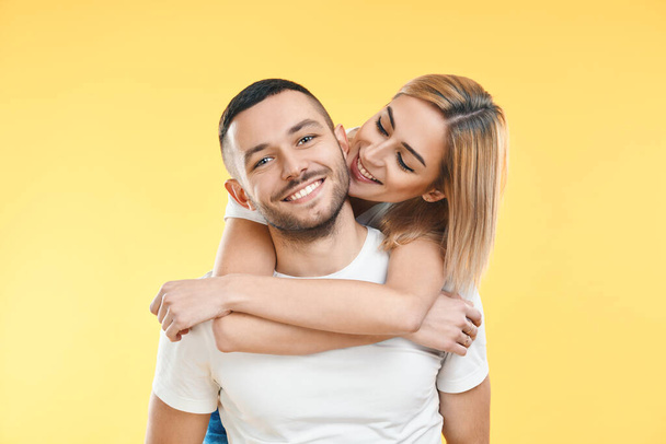 Happy smiling couple in love on yellow background. Man carrying his girlfriend piggyback. Love, romance, fun concept         - 写真・画像