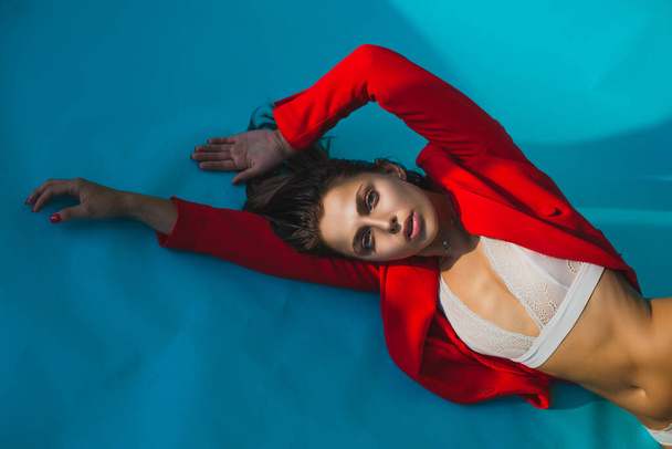 Beautiful woman lies in lace underwear on blue background in red jacket. Model posing lying on bright background with place for inscription - Photo, Image