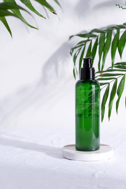 Botella de plástico verde con loción natural para una piel limpia sobre un fondo textil blanco con hojas tropicales verdes
. - Foto, Imagen