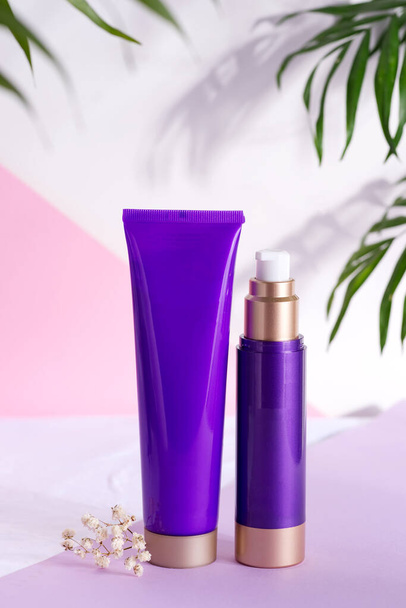 Paarse cosmetische plastic flessen voor crème en lotion met groene kruidenbladeren en bloemen op een driekleurige achtergrond. - Foto, afbeelding
