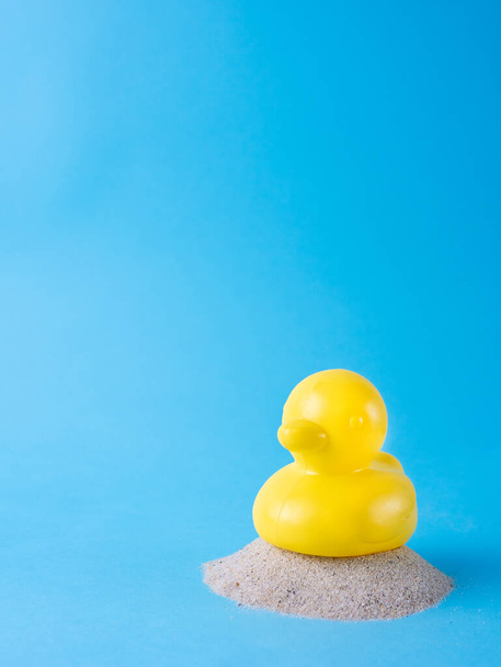 Gummi-gelbe Ente auf einer kleinen Menge Sand, isoliert auf blauem Hintergrund, mit Kopierraum, Sommerferienkonzept - Foto, Bild