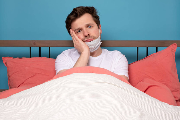 Gloomy european man with medical mask showing boredom while leaning head on hand and sitting alone at the bed. I feel so alone in quarantine concept. - Photo, Image