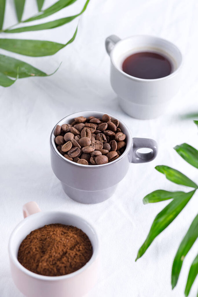 Ceramic cups with freshly roasted coffee beans and fresh ground aromatic coffee on a light background and green palm leaves. - Photo, Image
