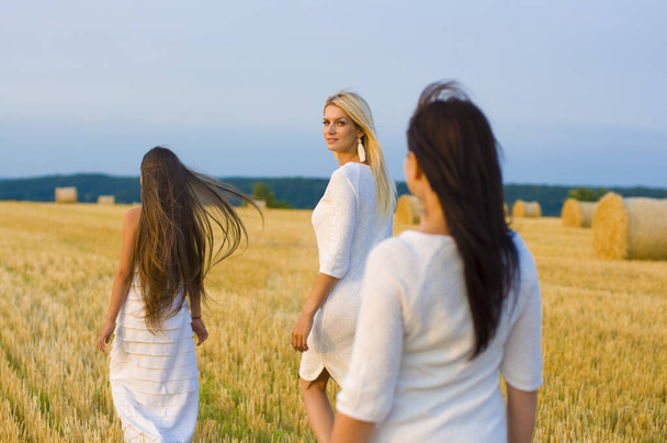een mooie vrouw van middelbare leeftijd en haar twee dochters, een blonde en een brunette, wandelen langs een tarweveld tegen de achtergrond van de prachtige zomerse natuur - Foto, afbeelding