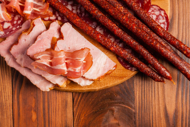 Assorted meat snacks on a wooden cutting board. Sausage, ham, bacon, smoked meats. Stock photo of meat products with blank space. - Fotó, kép