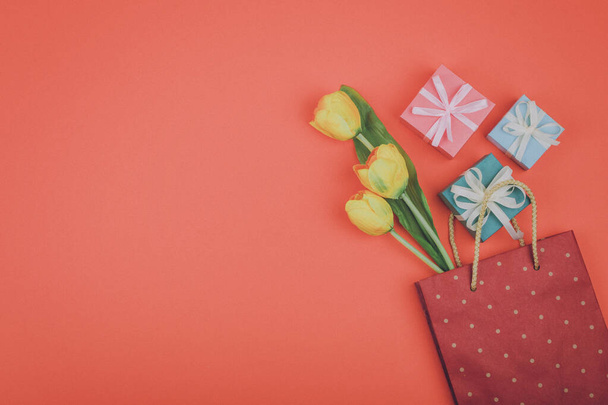 Flores de primavera y regalo en bolsa de papel sobre fondo rosa coral. Elegante piso tendido. Concepto festivo moderno mínimo
. - Foto, imagen