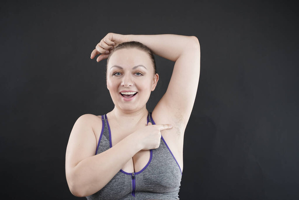 jong gelukkig plus size blank vrouw toont haar unshaved oksel op zwart studio achtergrond - Foto, afbeelding