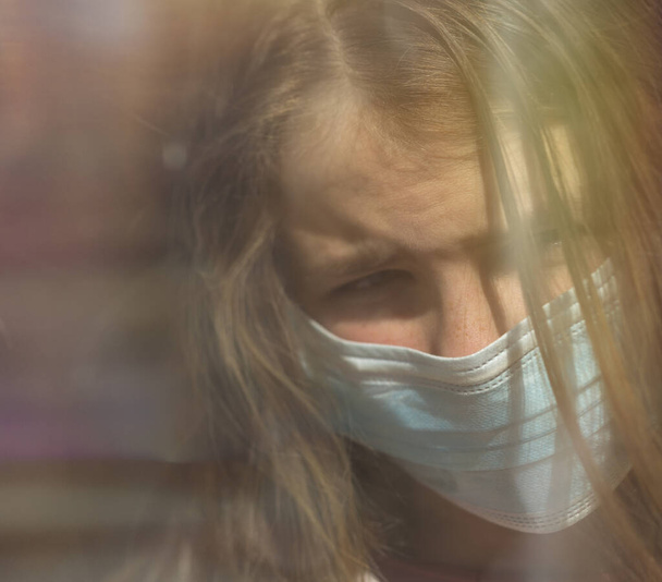 Sad  girl in protective medical mask looks out the window.Sick girl look at the window with longing on the street . Sadness child at home in isolatio - Fotografie, Obrázek