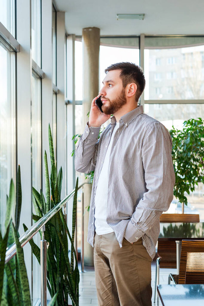 Phone conversation in urban modern office. Smart beard man is negotiating by smartphone. Attractive businessman freelancer in cafe. Young caucasian man holding smartphone for business work - Photo, Image