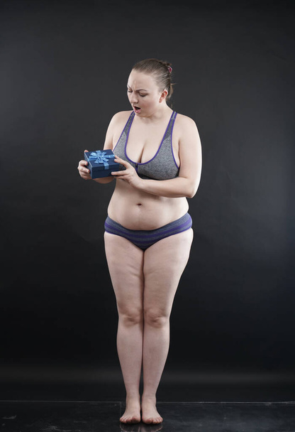 Beautiful plus size woman wearing sport underweaer holds box with gift subscription to a fitness on black studio solid background - Φωτογραφία, εικόνα
