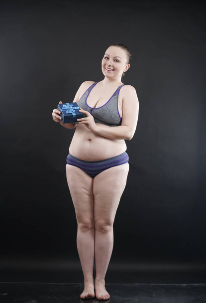 Schöne Plus-Size-Frau in Sport-Unterwäsche hält Schachtel mit Geschenk-Abonnement für ein Fitness-Studio auf schwarzem Studiohintergrund - Foto, Bild